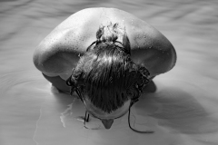 Surrender, Fine art black and white photograph of a woman immersed in calm water with her head lowered, creating a minimalist and emotional composition.