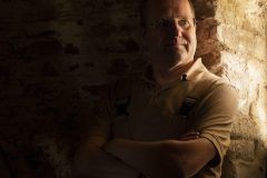 Portrait of a man with glasses looking toward window light in a dark rustic interior with warm side lighting.