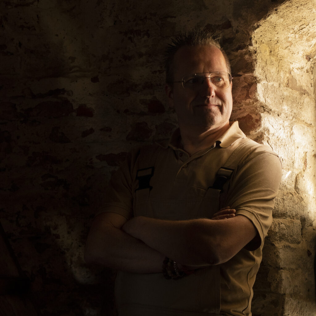 Portrait of a man with glasses looking toward window light in a dark rustic interior with warm side lighting.